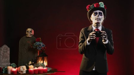 Téléchargez les photos : Femme avec dame de la mort corps art jouer à des jeux vidéo en studio, en utilisant contrôleur pour jouer sur la console de jeu. Portant fantasmagorique costume d'Halloween du jour mexicain de la tradition morte. - en image libre de droit