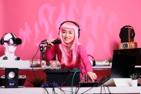 Photo for Artist with pink hair mixing techno song with electronic using professional turntables while enjoying talking with fans, having fun together during night time in club. Woman enjoying performing music - Royalty Free Image