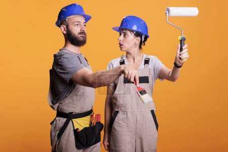 Photo for Young construction workers posing with painting tools, standing over yellow background. Team of constructors holding paintbrush and roller brush to work on refurbishment project in studio. - Royalty Free Image