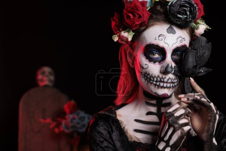Téléchargez les photos : Femme muerte fantasmagorique santa posant avec des roses noires, agissant comme une sainte déesse de la mort le jour de la fête mexicaine morte. Célébration du rituel d'Halloween avec la cavalera catrina crâne art corporel. - en image libre de droit