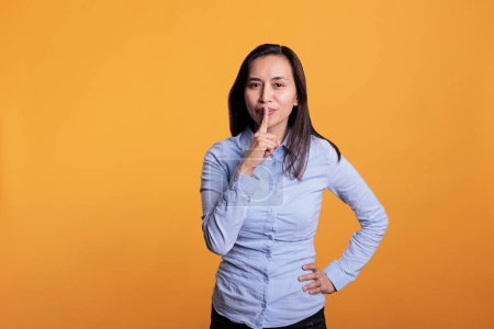 Téléchargez les photos : Portrait de femme philippine faisant un geste de silence en mettant l'index sur les lèvres posant sur fond jaune. Femme silencieuse qui fait taire les gens, ce qui témoigne du secret et de la confidentialité. - en image libre de droit
