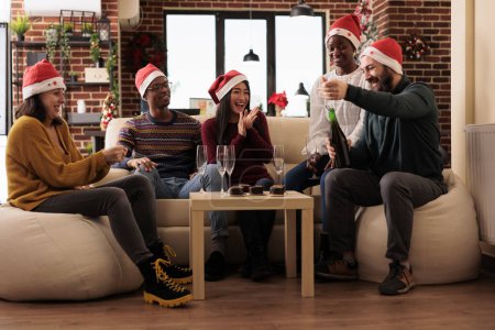 Foto de Compañeros alegres emocionados abriendo botella de vino espumoso en la fiesta de Navidad en la oficina decorada. Felices trabajadores reuniéndose alrededor de la mesa con alcohol y comida en el lugar de trabajo en víspera de Navidad - Imagen libre de derechos
