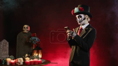 Photo for Creepy young woman browsing internet on smartphone, wearing suit and hat to celebrate mexican halloween day. Santa muerte model using online website on mobile phone, texting messages. - Royalty Free Image