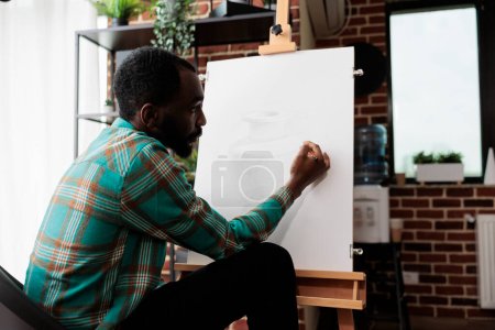 Téléchargez les photos : Jeune Afro-Américain s'exprimant à travers l'art, suivant des cours de dessin, étudiant masculin améliorant les compétences de croquis. Artiste talentueux assis au chevalet créer chef-d'œuvre sur toile - en image libre de droit