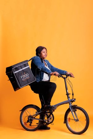 Téléchargez les photos : Travailleur pizzeria portant sac à dos à emporter alimentaire debout sur le vélo en studio avec fond jaune, livraison commande de pizza pour le dîner. Service de transport alimentaire et concept à emporter - en image libre de droit