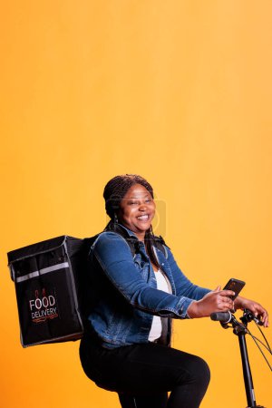 Téléchargez les photos : Coursier afro-américain à vélo tout en livrant des plats à emporter au client, vérifier adreess sur l'application de sortie de téléphone, debout sur fond jaune en studio. Service à emporter et concept - en image libre de droit