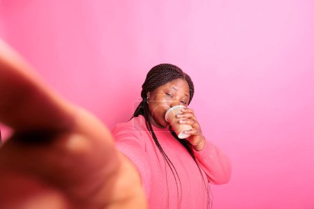 Téléchargez les photos : Joyeux modèle afro-américain tenant tasse à boire du café pendant la réunion de vidéocall en utilisant le téléphone tout en se tenant en studio sur fond rose. Femme ayant une émotion positive, appréciant le temps libre - en image libre de droit
