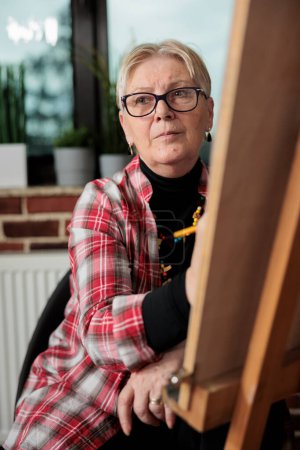 Photo for Art classes for seniors. Pensive mature woman sitting in front of easel looking aside, listening to teacher during drawing lesson, learning sketching in small groups. Older people and creativity - Royalty Free Image