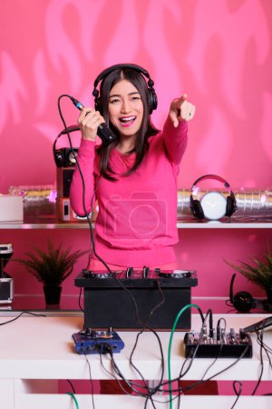 Photo for Asian artist performing electronic music using professional turntables, enjoying to perform in club at night. Smiling performer mixing techno sound, talking into microphone with fans - Royalty Free Image