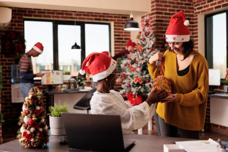 Photo for Smiling company woman worker giving festive bag with present to african american coworker at christmas eve. Employee working on laptop and receiving xmas gift from caucasian colleague - Royalty Free Image