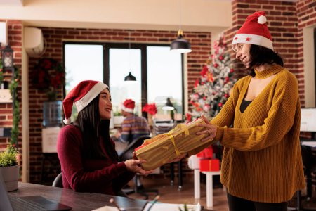 Photo for Two diverse women colleagues sharing christmas gift while working on laptop in decorated office. Smiling startup company employee greeting coworker and giving xmas present - Royalty Free Image