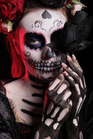 Photo for Beautiful lady of dead holding black roses in studio, looking like la cavalera catrina skull with make up. Santa muerte goddess with traditional halloween costume on dios de los muertos. - Royalty Free Image