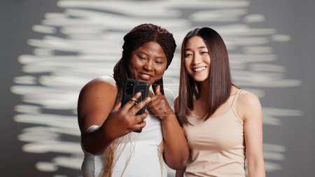 Téléchargez les photos : Interracial cheerful ladies taking photos with phone, female models promoting different skintones and body types. Confident natural women laughing with pictures for skincare campaign. - en image libre de droit