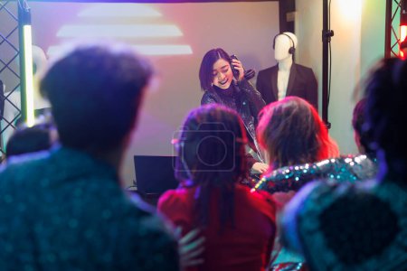 Photo for Crowd watching musician performance while attending discotheque party in nightclub. Young asian woman dj playing using headphones and digital controller on stage with spotlights in club - Royalty Free Image