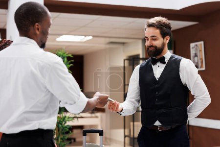Photo for Hotel porter taking money from client, accepting to carry luggage and help with bags after check in process. Male steward working as bellboy at luxury hotel, receiving cash tip from guests. - Royalty Free Image