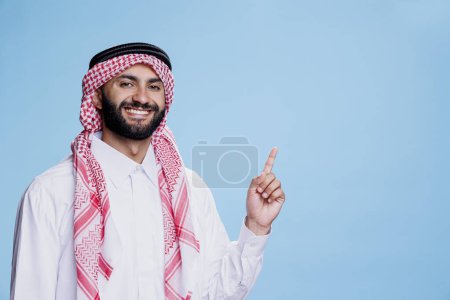 Téléchargez les photos : Homme musulman joyeux vêtu d'une robe traditionnelle pointant vers le haut avec une expression souriante. Produit publicitaire arabe insouciant tout en montrant dans la direction vers le haut et en regardant la caméra - en image libre de droit