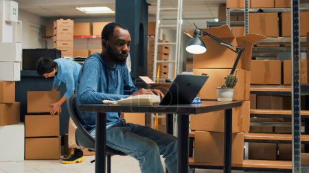 Téléchargez les photos : African american worker planning logistics to ship products, preparing order shipment with stock inventory information. Male owner working on laptop with packages from warehouse racks. - en image libre de droit