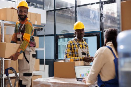 Photo for Warehouse package handlers and order picker working in storage room. Storehouse workers wearing safety hard hats taking cardboard boxes from shelf while manager checking products list - Royalty Free Image
