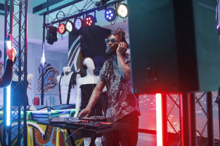 Foto de Dj en auriculares mezclando sonidos usando un controlador digital de música electrónica en el escenario en la discoteca. Joven afroamericano músico hombre que actúa en la fiesta discoteca en el club - Imagen libre de derechos