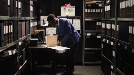 Photo for Professionally dressed african american investigator examines confidential evidence in organized office space. Private detective holding certain files from shelves for study and forensic analysis. - Royalty Free Image