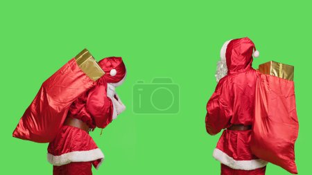 Téléchargez les photos : Jeune homme dépeignant Santa Claus sur fond d'écran vert, se préparant à livrer des cadeaux aux enfants du monde entier. Père Noël avec un énorme sac rouge de jouets, saison décembre. - en image libre de droit