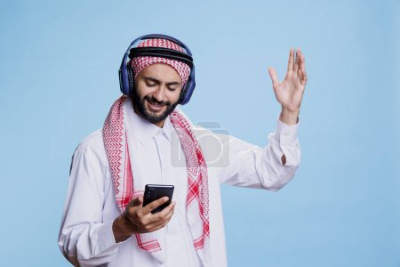 Téléchargez les photos : Homme musulman portant un foulard ghutra et écouteurs sans fil changer de chanson dans la liste de lecture sur smartphone. Écouter de la musique arabe dans les écouteurs, gesticuler et utiliser un téléphone portable - en image libre de droit