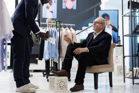 Foto de Trabajador afroamericano ayudando a clientes mayores con zapatos de moda en el centro comercial. Hombre mayor comprando ropa elegante, analizando la colección de moda en boutique. Concepto de moda - Imagen libre de derechos