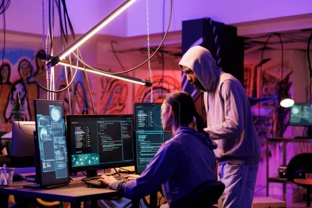 Hackers coding malicious software on computer together while working in abandoned warehouse. Internet thieves in hoods planning phishing attack and programming malware in dark room