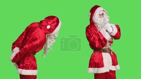 Foto de Hombre de traje rojo comprueba reloj de pulsera para ver el tiempo, posando sobre fondo de pantalla verde. Persona segura de usar traje festivo de Santa Claus para difundir el espíritu navideño, celebrar las vacaciones de invierno. - Imagen libre de derechos