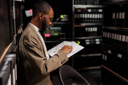 Photo for Investigator studying folder with police records in archive in office. African american detective standing near shelf, working late and analyzing forensic reports at night time - Royalty Free Image
