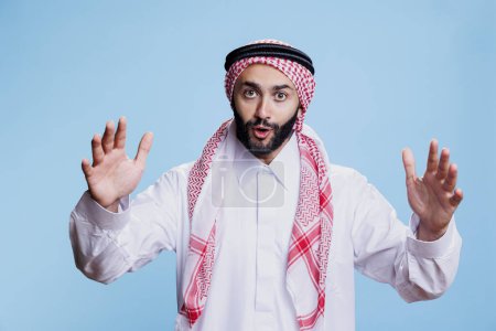 Foto de Hombre excitado vestido con ropa tradicional musulmana levantando las manos y hablando retrato de estudio. Persona que usa pañuelo árabe para la cabeza y que gesticula mientras habla y mira la cámara - Imagen libre de derechos