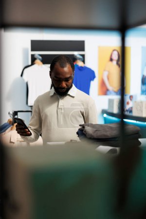 Téléchargez les photos : Homme afro-américain vérifiant le prix des vêtements sur le site Web en ligne sur smartphone tout en choisissant la tenue dans le magasin de vêtements. Client utilisant un téléphone portable tout en sélectionnant le vêtement dans le centre commercial - en image libre de droit