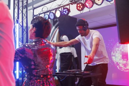 Foto de El joven DJ mezclando pistas de techno en el escenario mientras la gente festejaba en la pista de baile en el club nocturno. Músico tocando música electrónica en el escenario mientras los clubbers bailan en la discoteca - Imagen libre de derechos