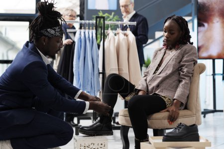 Téléchargez les photos : Travailleur Showroom aider le client afro-américain avec l'achat dans le centre commercial. Client élégant essayant des chaussures élégantes, l'achat de marchandises à la mode dans la boutique moderne. Concept d'activité commerciale - en image libre de droit
