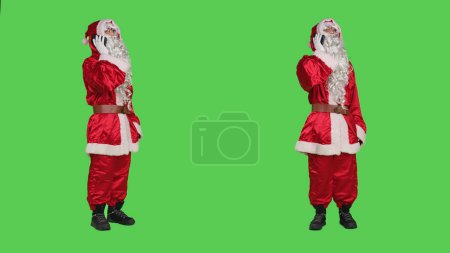 Photo for Saint nick answers phone call in studio, using smartphone mobile telephone line to have remote conversation. Christmas character talking to people against full body greenscreen. - Royalty Free Image