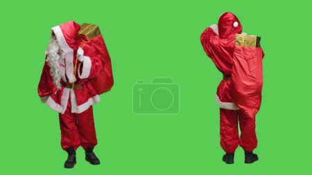 Photo for Father christmas looks around in studio, carrying big sack filled with presents boxes over full body greenscreen. Young man dressed as santa spreading xmas holiday spirit, having gifts for kids. - Royalty Free Image