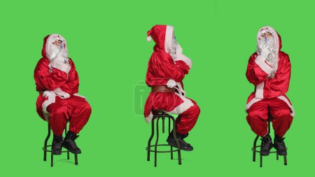 Foto de Personaje en traje rojo sentarse en la silla, pensando en nuevas ideas sobre fondo de pantalla verde de cuerpo completo. Joven hombre pensativo actuando como Santa Claus en traje y barba blanca, concepto de lluvia de ideas. - Imagen libre de derechos