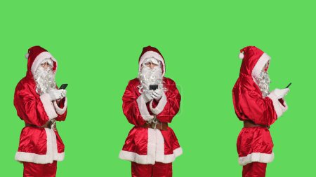 Téléchargez les photos : Père Noël à la recherche d'idées cadeaux sur smartphone, en utilisant le navigateur en ligne pour diffuser la positivité de Noël. Santa claus en costume tenant mobile pohne sur fond vert, événement de vacances. - en image libre de droit