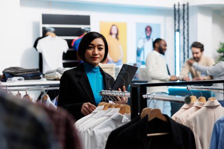 Photo for Clothing store asian woman manager using digital tablet to examine apparel inventory and looking at camera. Shopping center young employee standing near garment rack portrait - Royalty Free Image