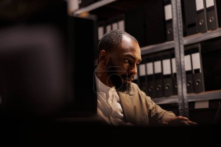 Téléchargez les photos : Un flic afro-américain mène une enquête dans les archives de la police, assis près d'une étagère remplie de dossiers. Détective privé concentré résolvant une affaire de crime complexe - en image libre de droit