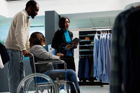 Photo for African american woman with chronic impairment getting advice from clothing store assistant while choosing outfit. Mall boutique customer in wheelchair shopping for casual apparel - Royalty Free Image