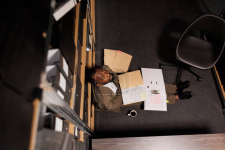 Téléchargez les photos : Un flic épuisé assis sur le sol entouré de papiers du dossier criminel. Enquêteur fatigué surmené étudiant la documentation policière au bureau tard et regardant vers le haut vue vers le haut - en image libre de droit
