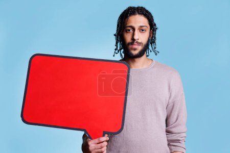 Téléchargez les photos : Homme arabe tenant bannière vide rouge avec place pour le texte pour la publicité mock up et en regardant la caméra. Modèle debout avec cadre de dialogue vide pour message avec portrait de l'espace de copie - en image libre de droit