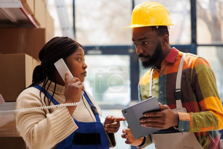 Photo for Storehouse manager answering logistics department call and giving stock information. African american warehouse worker showing report on tablet and supervisor discussing data on telephone - Royalty Free Image