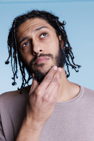 Photo for Arab young adult man rubbing chin in contemplation with puzzled facial expression. Attractive model with black braids hairstyle looking away while thinking with pensive emotions - Royalty Free Image