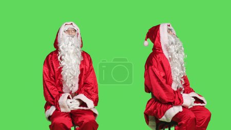Photo for Man dressed as santa claus on chair sitting against greenscreen background, father christmas embodiment. Young adult advertising december seasonal holiday, traditional event. - Royalty Free Image