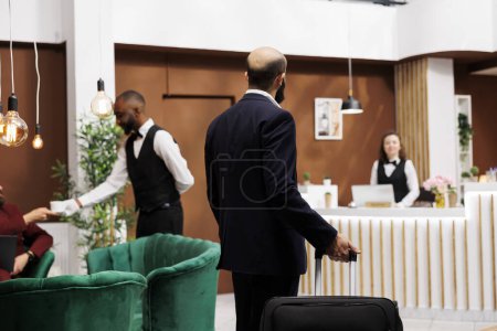 Photo for Businessman in suit arrive at hotel, entering reception lobby with suitcase luggage for international corporate meetings. White collar worker with baggage travelling for work, business trip. - Royalty Free Image