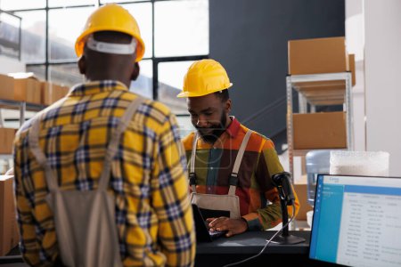 Photo for African american warehouse operators checking delivery schedule on laptop while standing at counter desk. Storehouse workers wearing overalls and helmets checking electronic orders list - Royalty Free Image