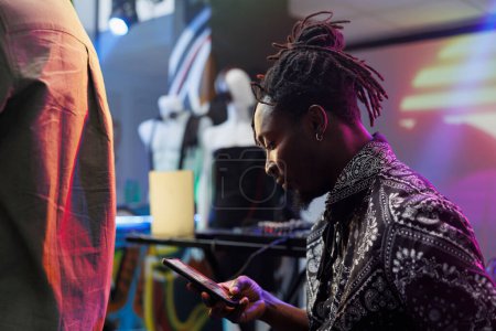 Young african american man using smartphone while resting in nightclub. Clubber scrolling social media and online pages on mobile phone while relaxing at discotheque party in club