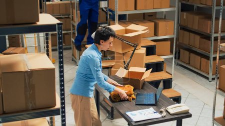 Téléchargez les photos : Woman employee taking supplies from warehouse shelves and putting them in cardboard boxes, using packages for delivery. Young adult writing inventory information on laptop, logistics. - en image libre de droit